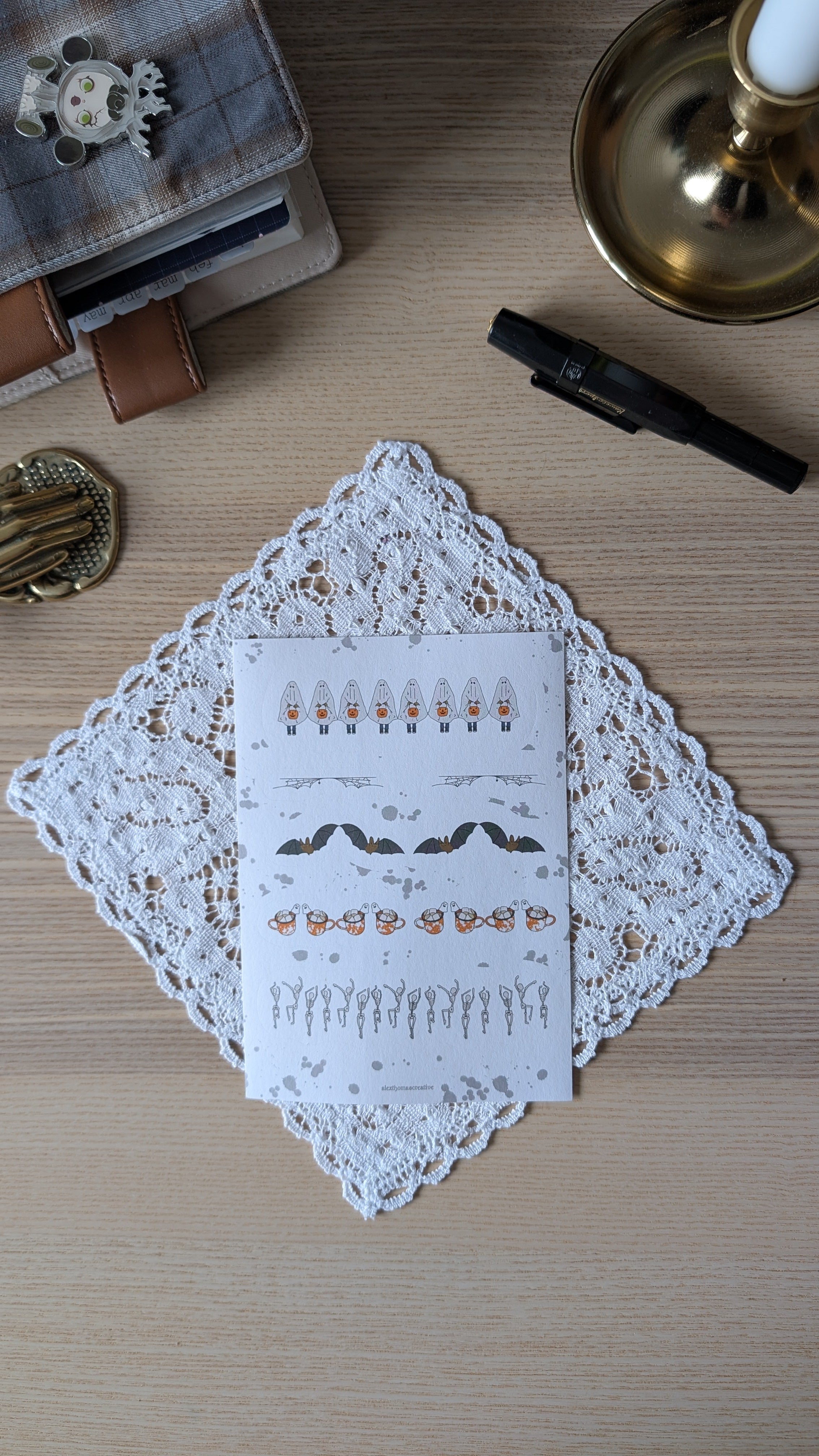 Sticker sheet featuring Halloween motifs such as ghosts, bats and skeletons. The sheet is sitting on a white doily on a wooden surface.