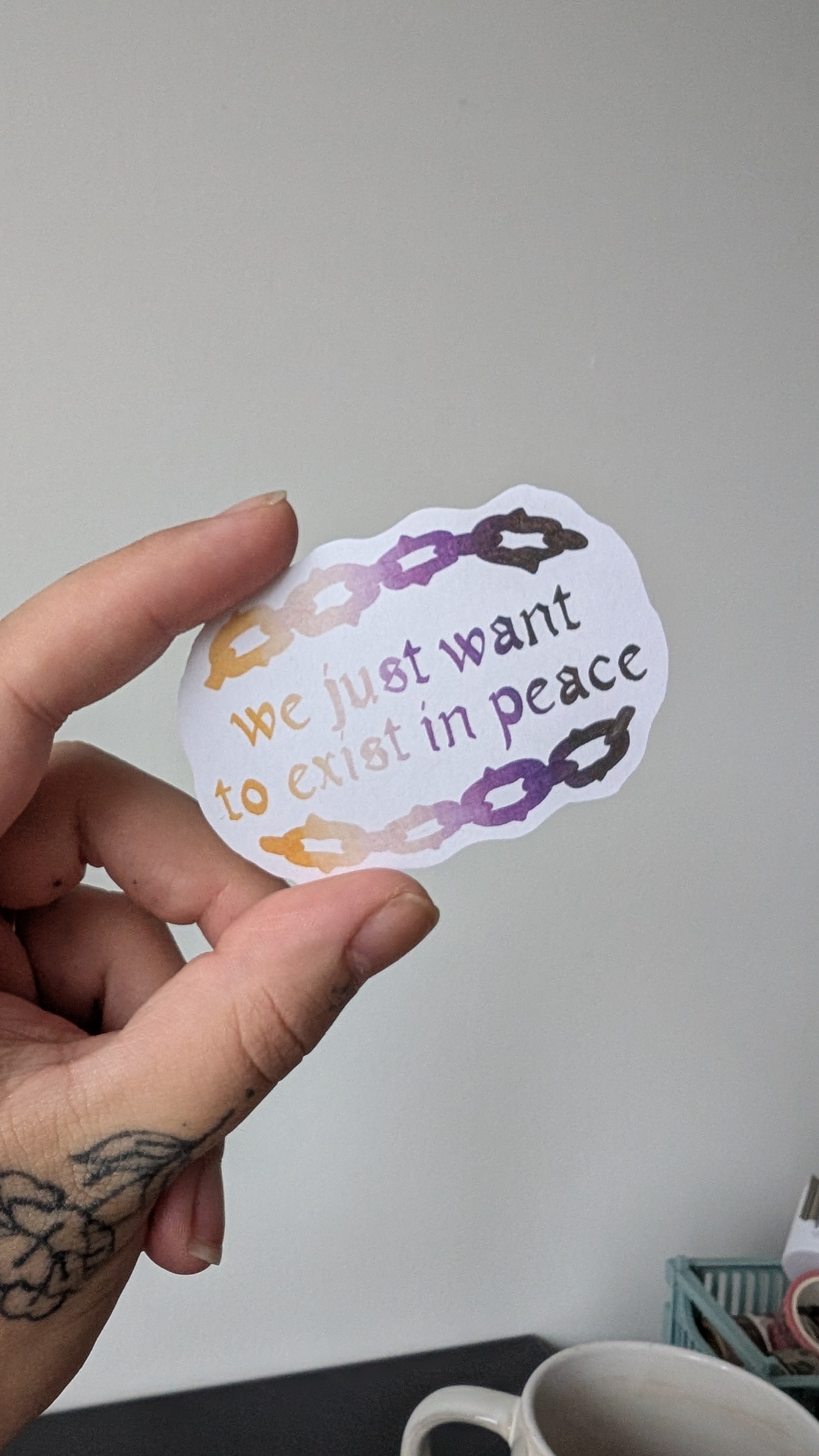 Sticker of text stating "we just want to exist in peace" with an illustrated chainlink border. The text and illustration on the sticker is in the colours black, purple, white and yellow (the colours of the non binary pride flag).