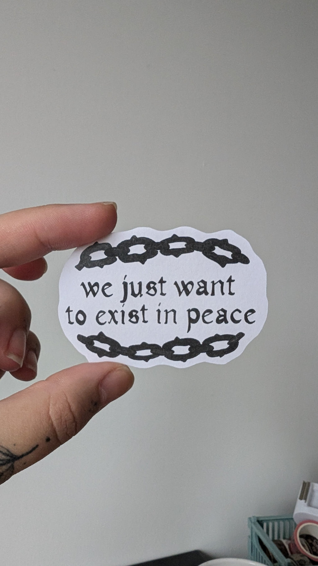 Sticker of text stating "we just want to exist in peace" with an illustrated chainlink border. The text and illustration on the sticker is in black.
