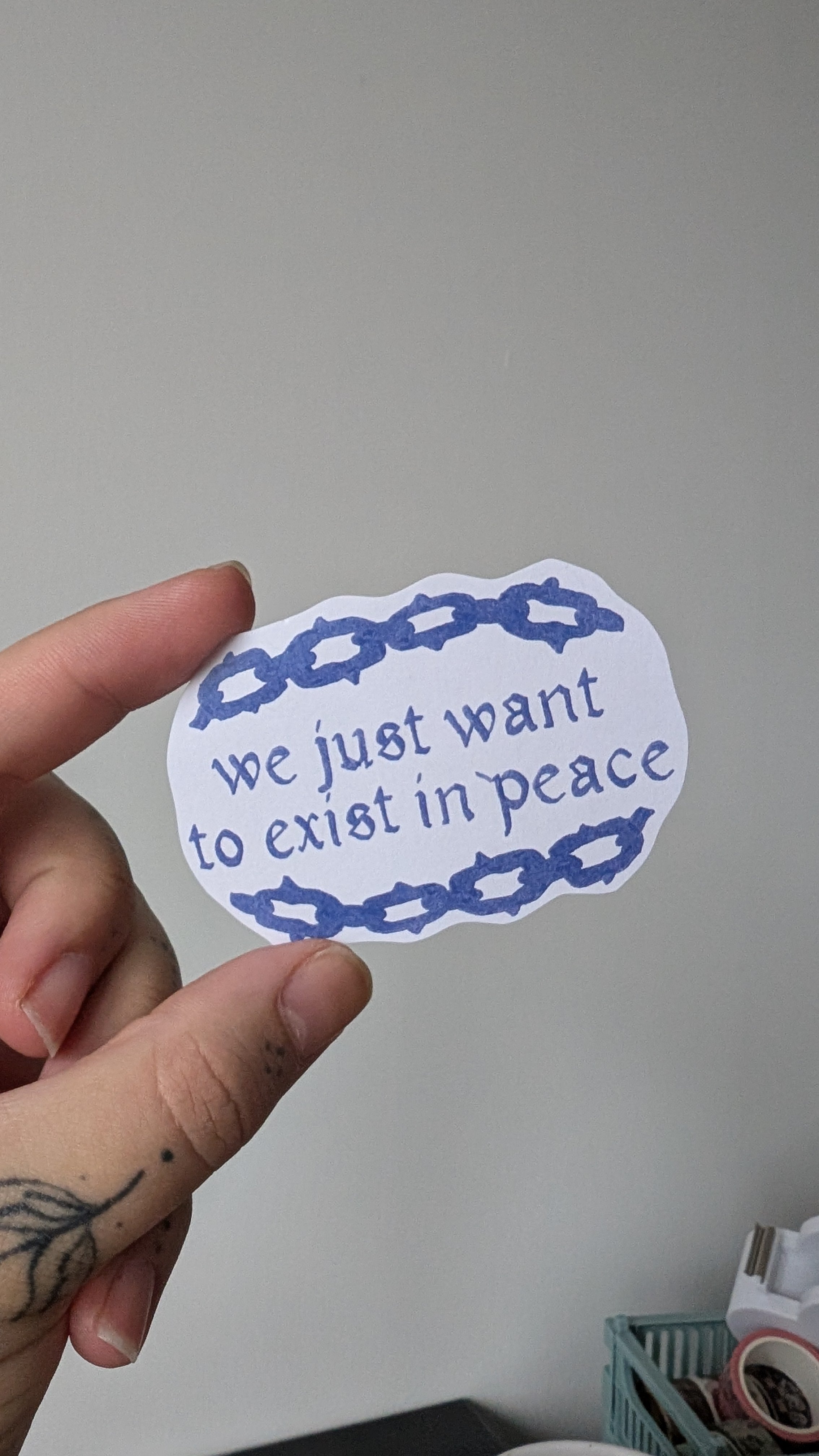 Sticker of text stating "we just want to exist in peace" with an illustrated chainlink border. The text and illustration on the sticker is in the colours blue.