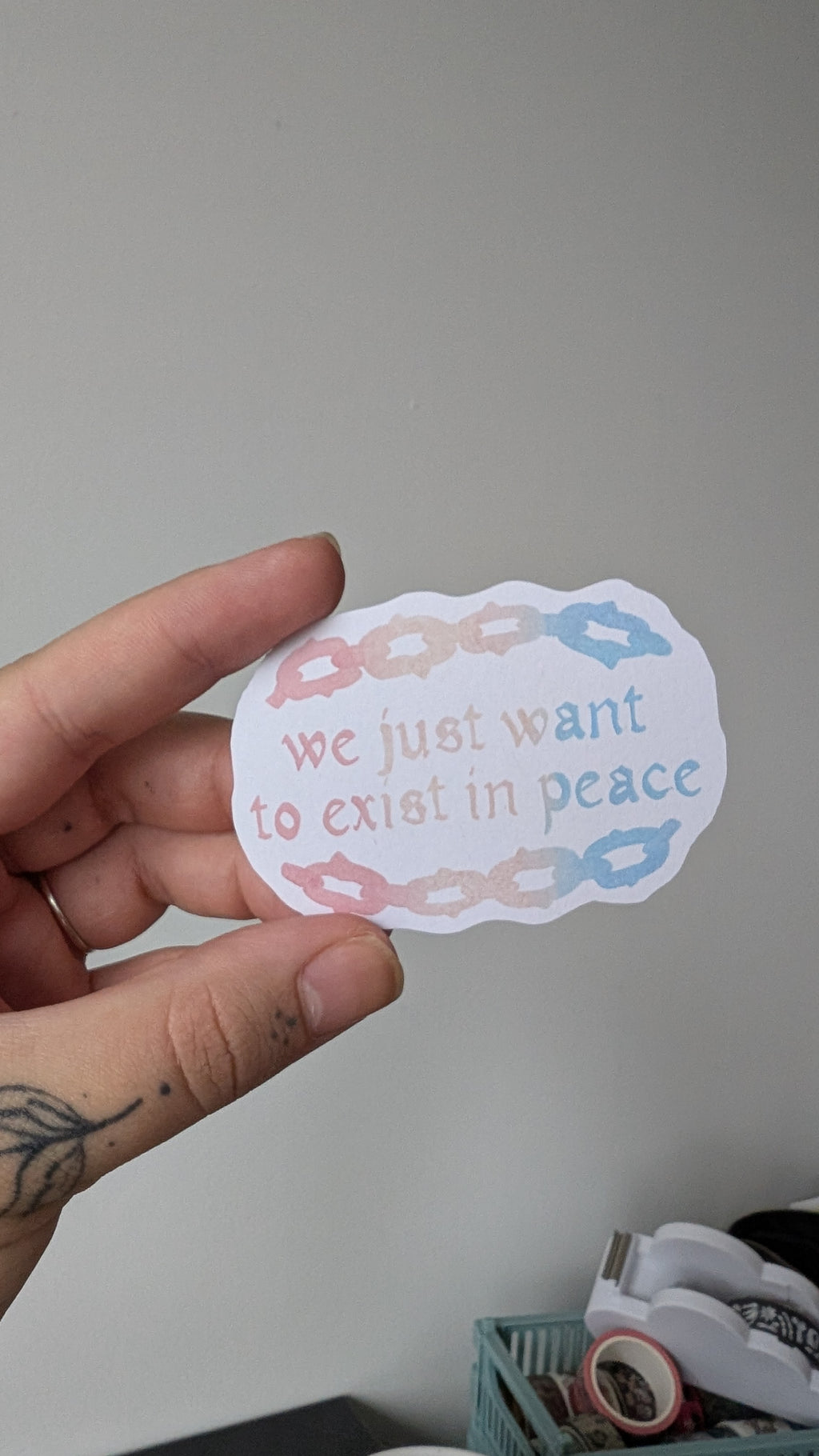 Sticker of text stating "we just want to exist in peace" with an illustrated chainlink border. The text and illustration on the sticker is in the colours blue, white and pink (the colours of the transgender pride flag).