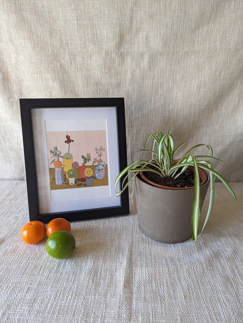 Art print of a selection of citrus fruits, atop vessels, with decorative flowers stuck into the fruit.  The print is photographed in a black frame, with a sheet behind it. A spider plant, some satsumas and a life sit beside the frame.