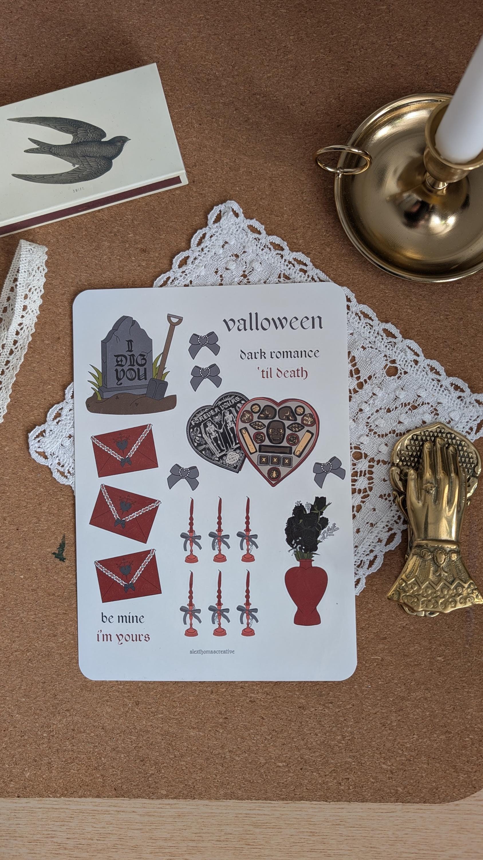Sticker sheet featuring Halloween and Valentine's Day motifs such as love letters (black) roses and a box of chocolates. The sheet is sitting on a white doily on a wooden surface.
