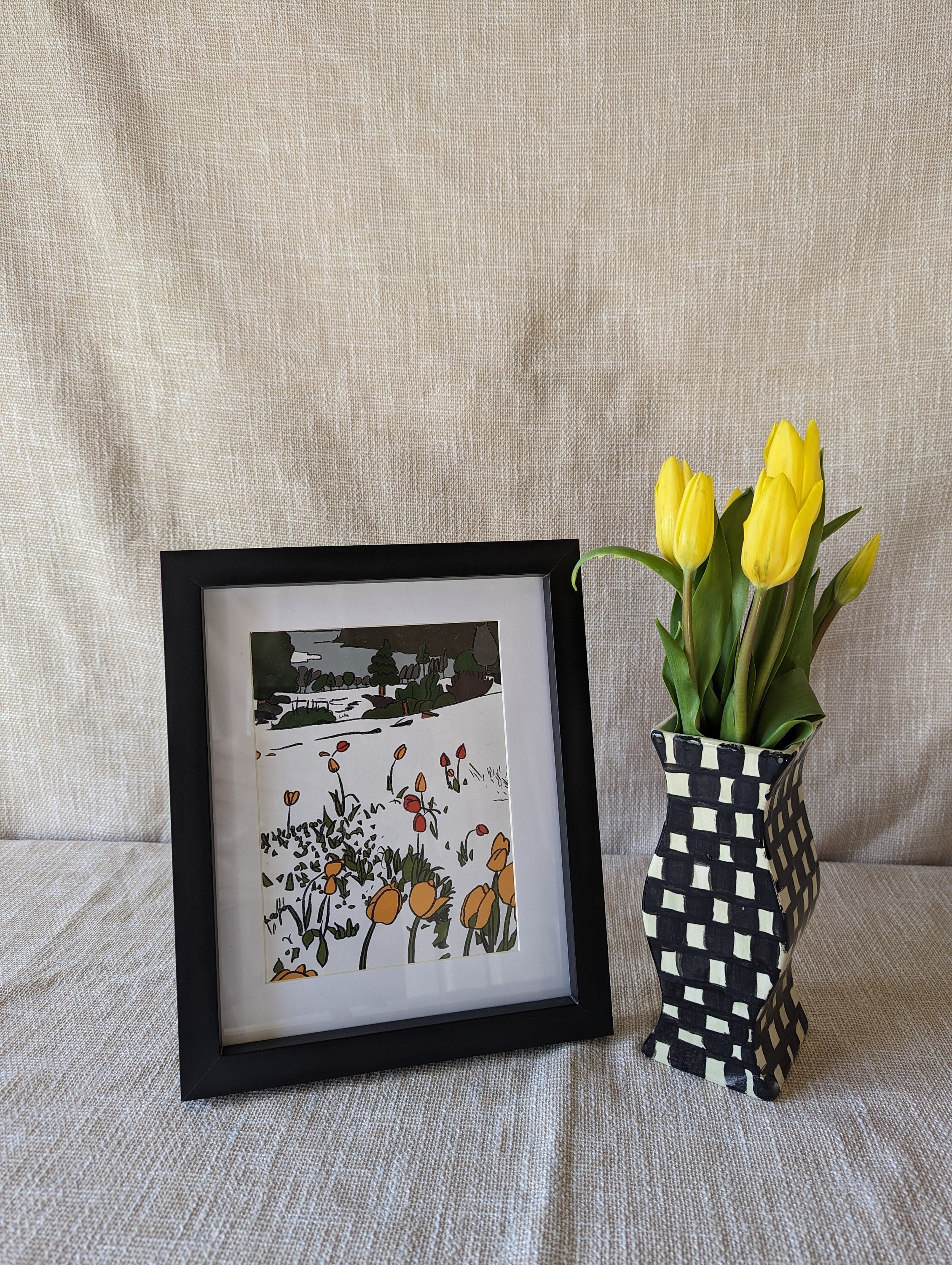 Art print of a snowy landscape. In the foreground and midground tulips sprout up through the snow. In the background, there are bushes, trees and a body of water. The print is photographed in a black frame, with a sheet behind it and a vase of tulips next to the frame.