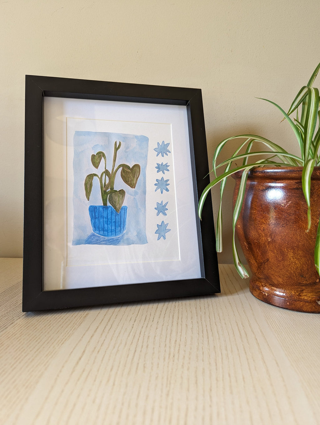 Art print of a monstera plant. The print is photographed in a black frame, with a sheet behind it and spider plant sits beside the frame.