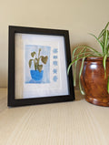 Art print of a monstera plant. The print is photographed in a black frame, with a sheet behind it and spider plant sits beside the frame.